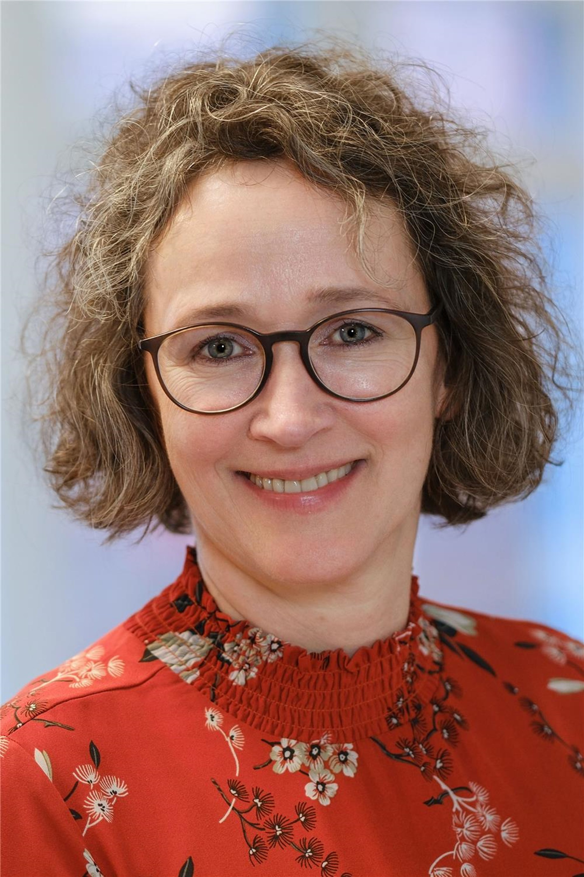 Kathrin Klein lächelt in die Kamera. Sie trägt eine braune Brille, kurzes lockiges Haar und eine geblümte rote, hochgeschlossene Bluse.