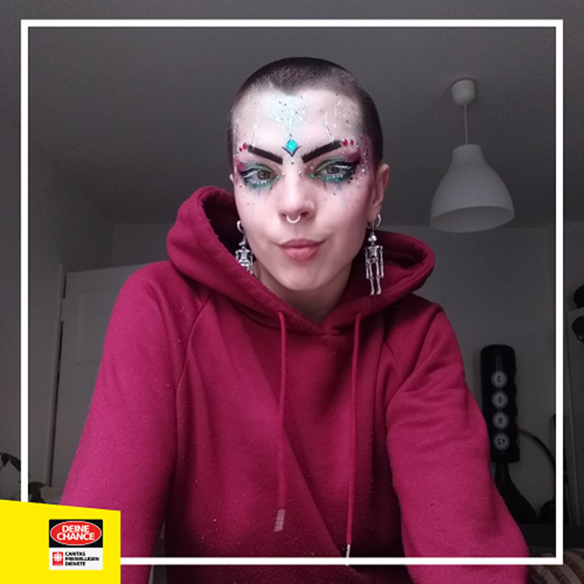 Sam trägt einen roten Pullover und große Ohrringe, ist stark geschminkt und hat viele Piercings und Tattoos im Gesicht. 