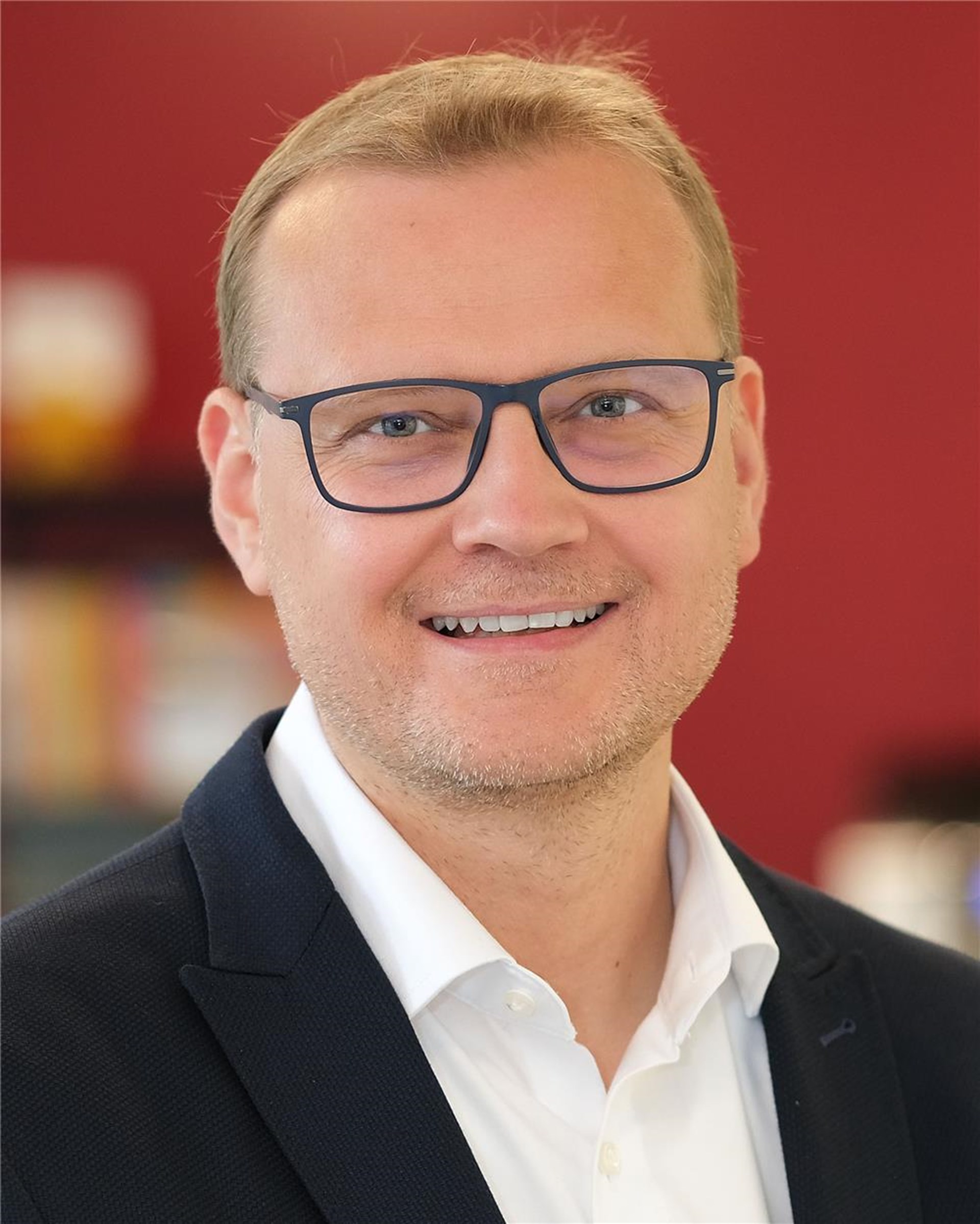 Lars Kohlenberg trägt eine schwarze Brille und lächelt in die Kamera. Er trägt ein weißes Hemd und ein schwarzes Sakko. Im Hintergrund eine verschwommene rote Wand.